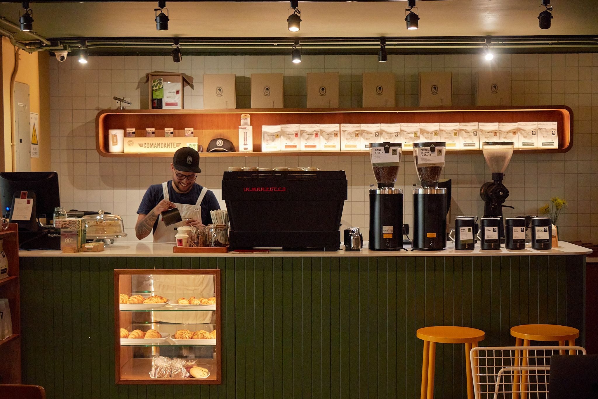 Barra de café con barista, vitrina de pastelería y butacos amarillos, Campesino Coffee, Medellín